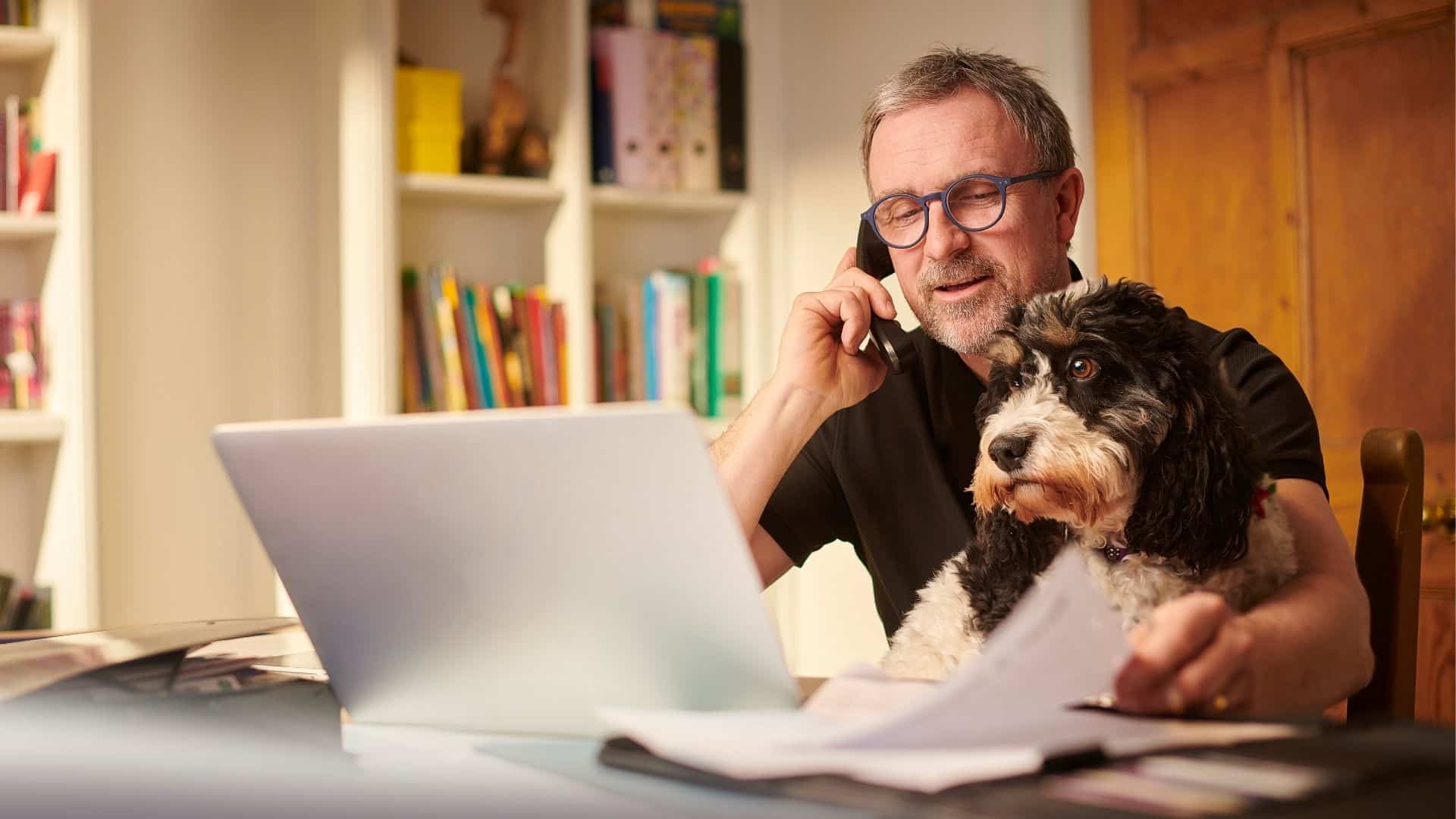 A man sat at his desk with his dog on his lap. He is on the phone and looking at his laptop screen, surrounded by papers.