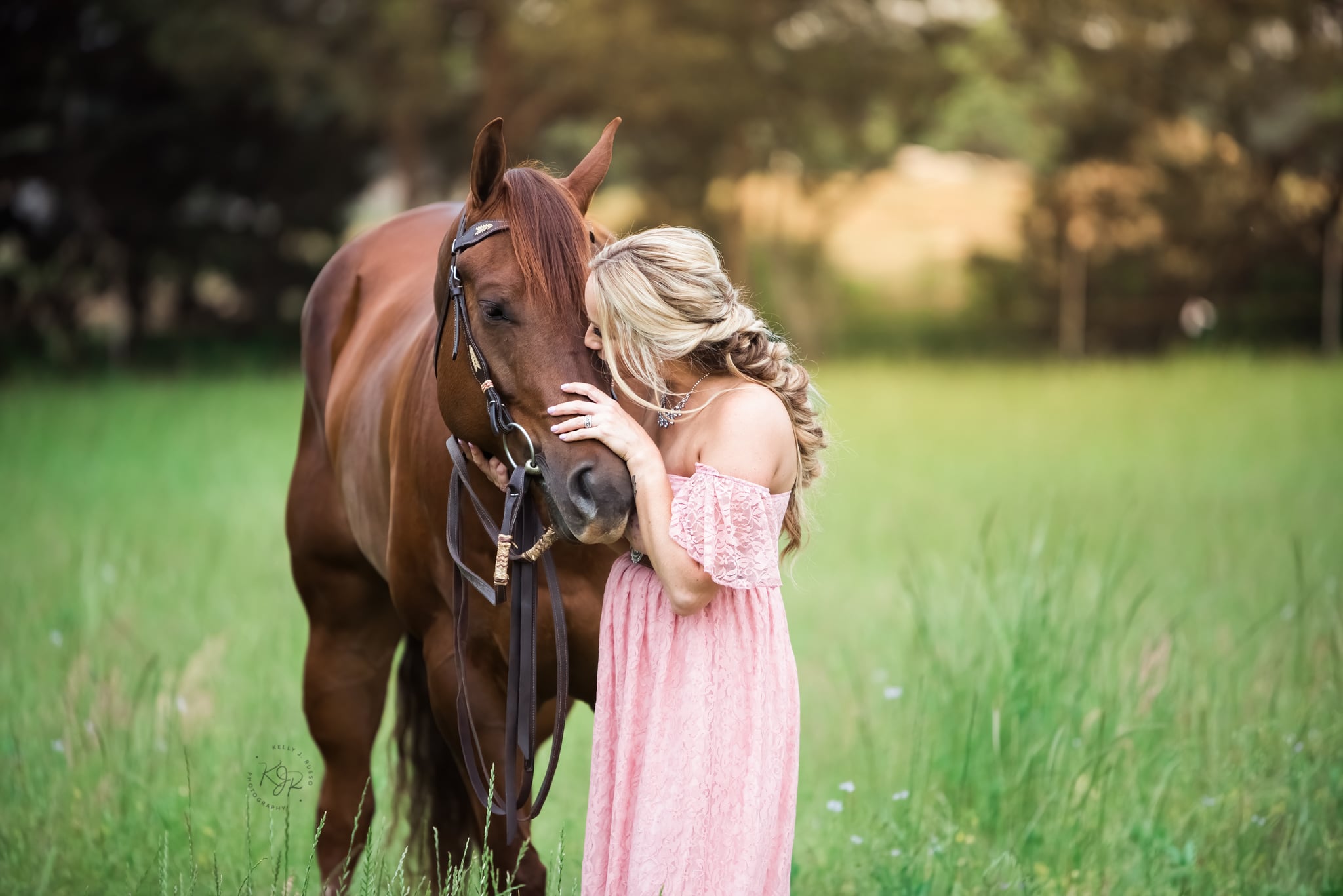 A professional photograph captured by Kelly J. Russo Photograpy showing a brown horse stood in a field with it owner, a blonder female in a pink dress, hugging it. 